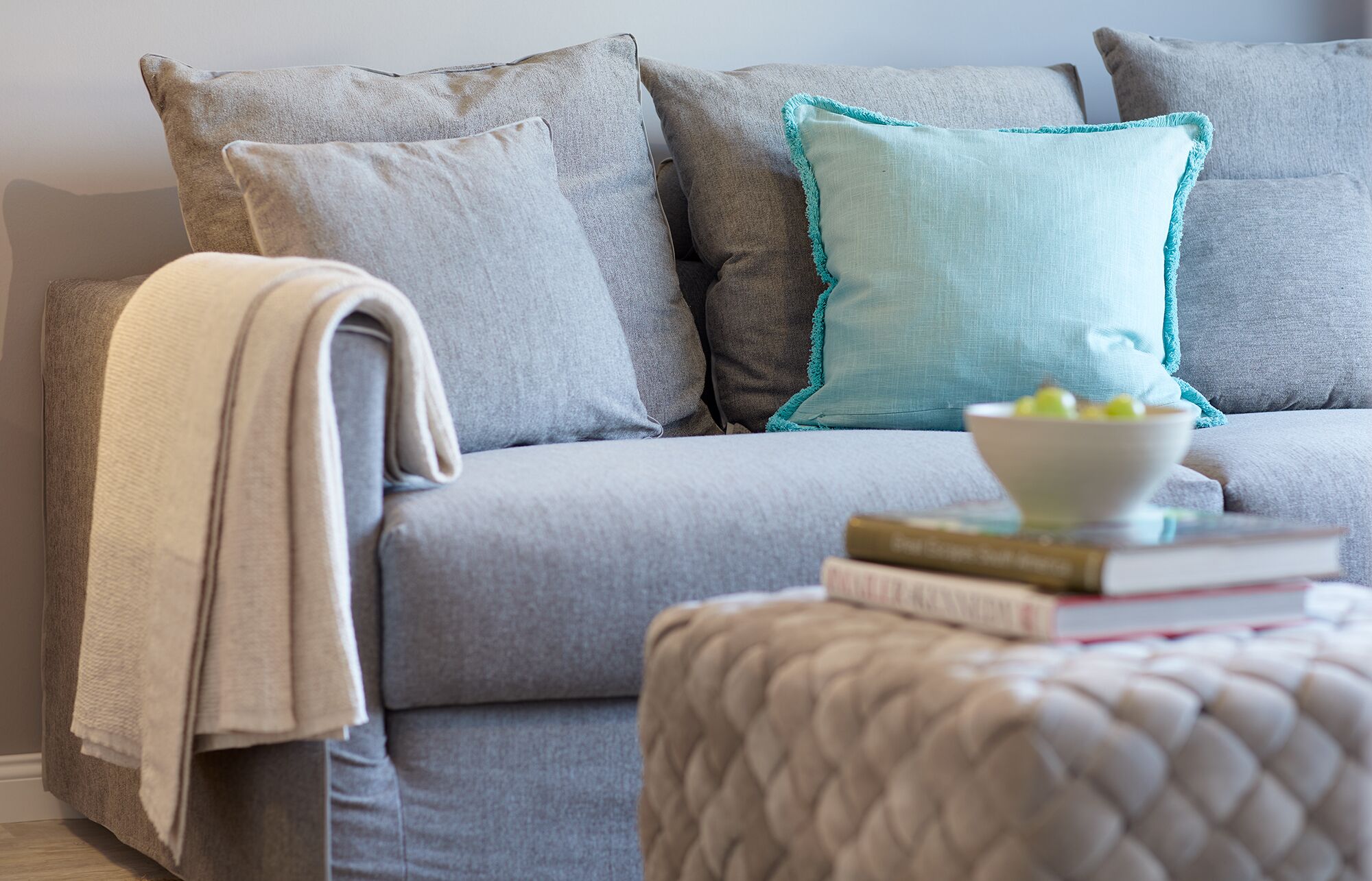 A grey sofa with multiple cushions, a white bowl with fruits, and a few books on a small ottoman.
