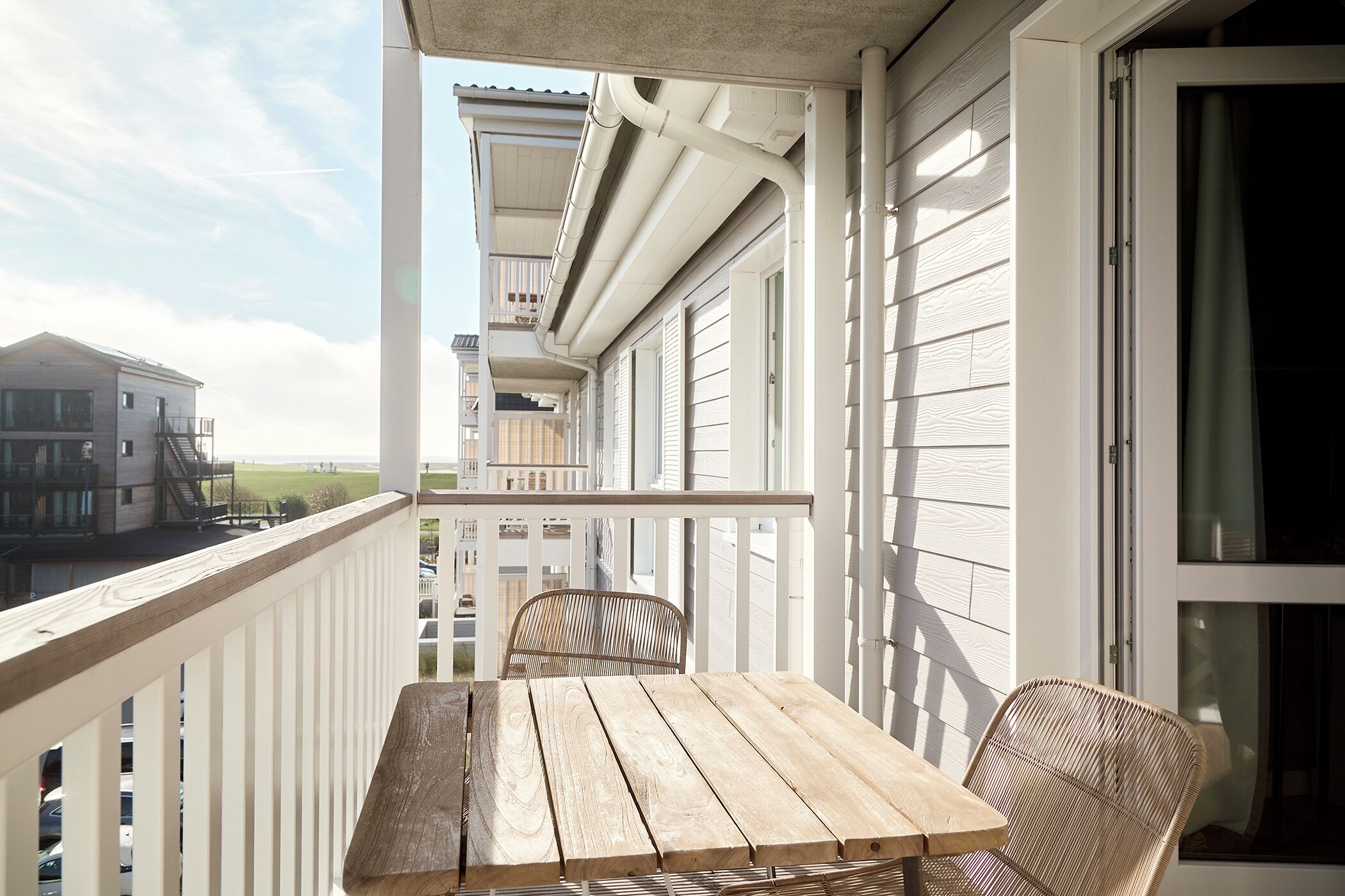 A wooden table and wicker chair on a balcony with white railings, overlooking a residential area.