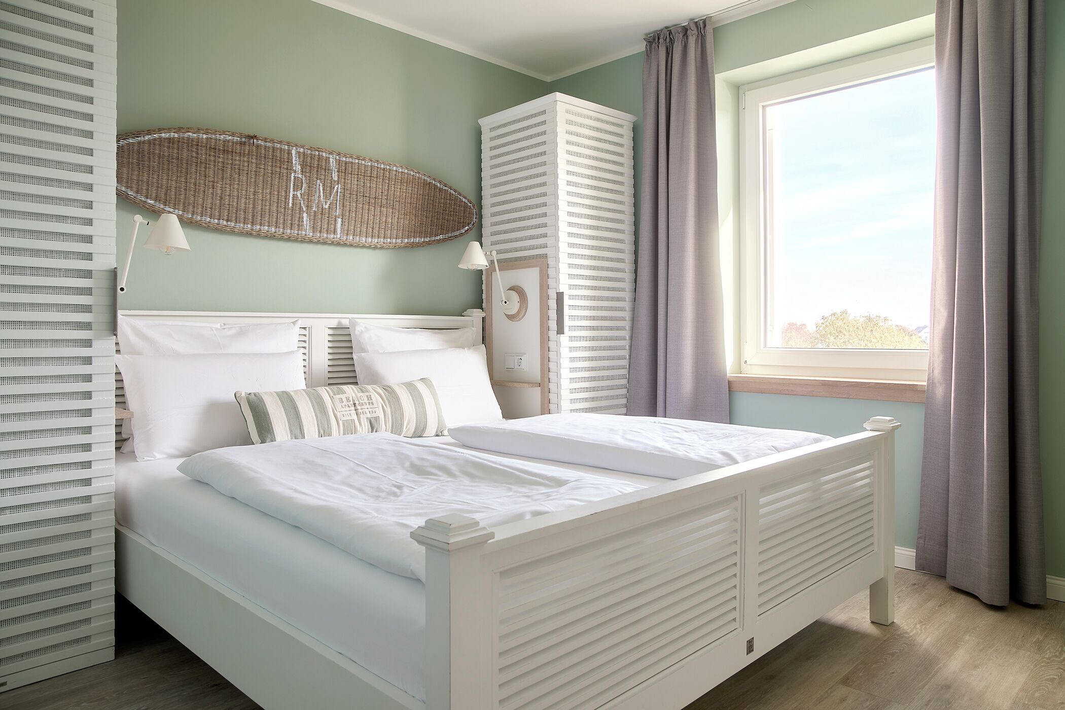 A neatly made bed with white bedding, a white headboard, and a bedside table with a lamp. There is a large window with curtains, and a tall white wardrobe against the wall.