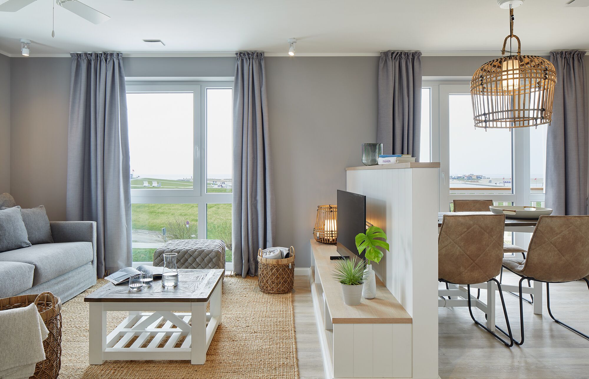 Een moderne woonkamer met een bank, salontafel en een eethoek. Er zijn grote ramen met gordijnen, een televisie en een hanglamp.