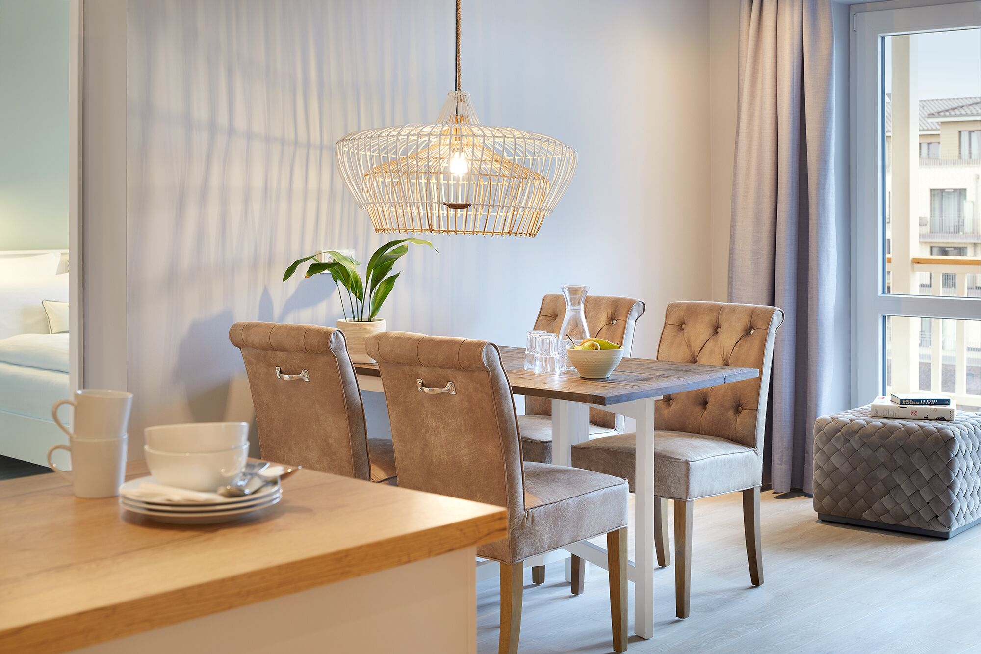 A modern dining area with a wooden table, four upholstered chairs, a hanging light fixture, a potted plant, and a view of the city through a window.