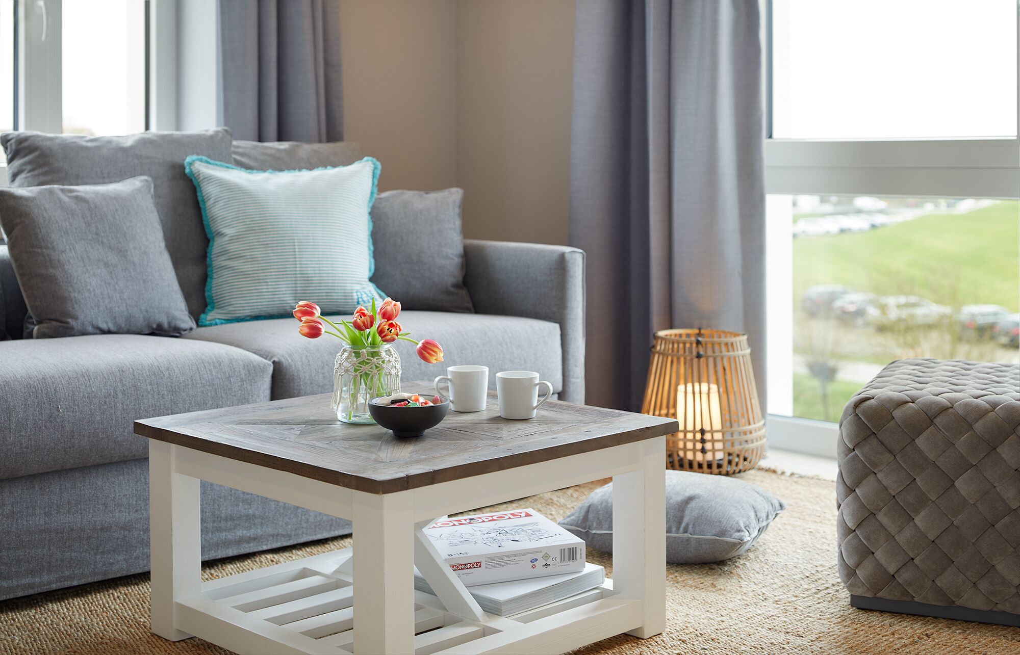 Un salon avec un canapé gris, une table basse sur laquelle se trouve un magazine, un vase rempli de fleurs et une vue sur l’extérieur.