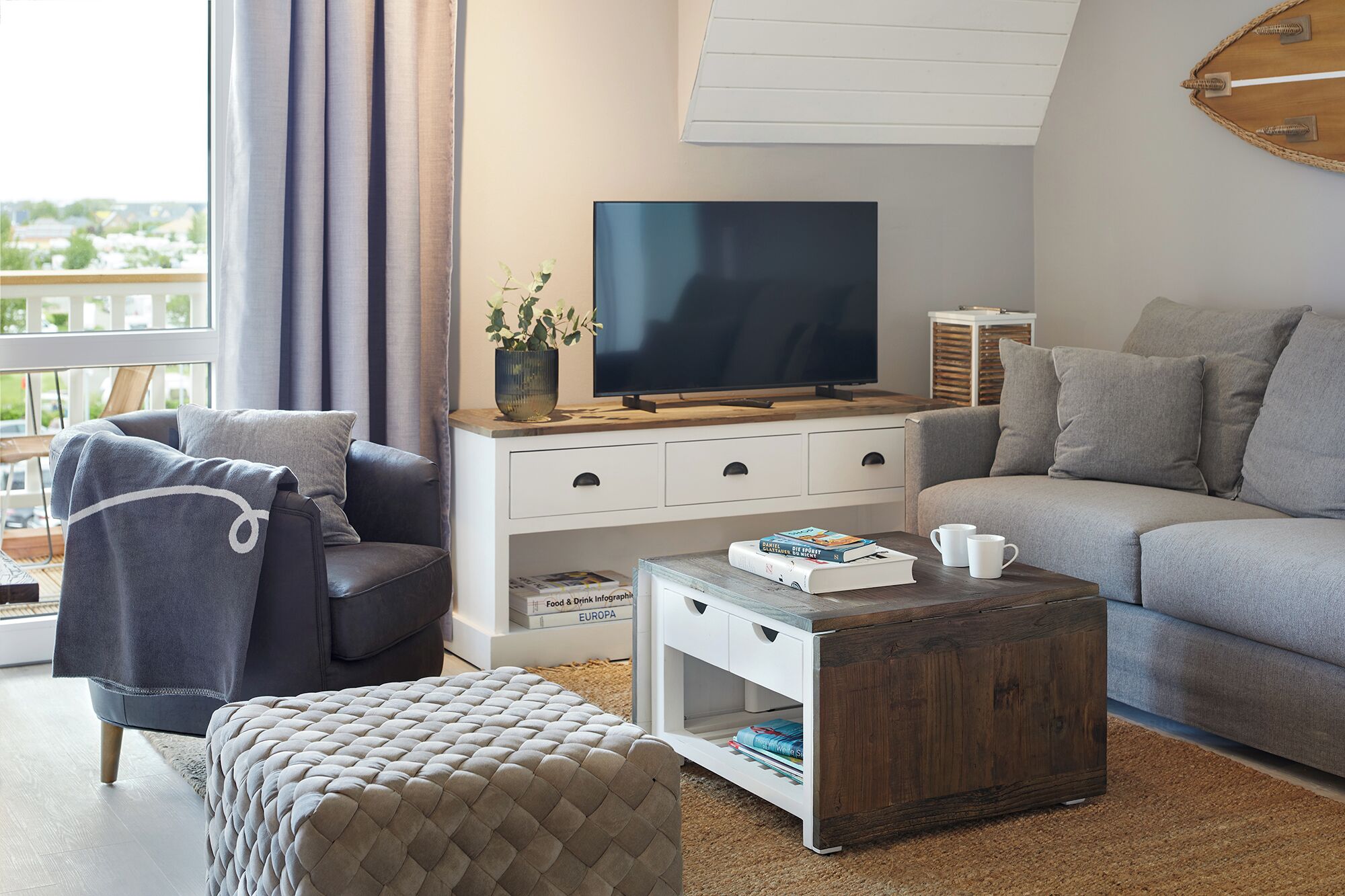 Ett modernt vardagsrum med en grå soffa, en tillhörande fåtölj, ett soffbord i trä och en platt-TV placerad på ett vitt skåp.