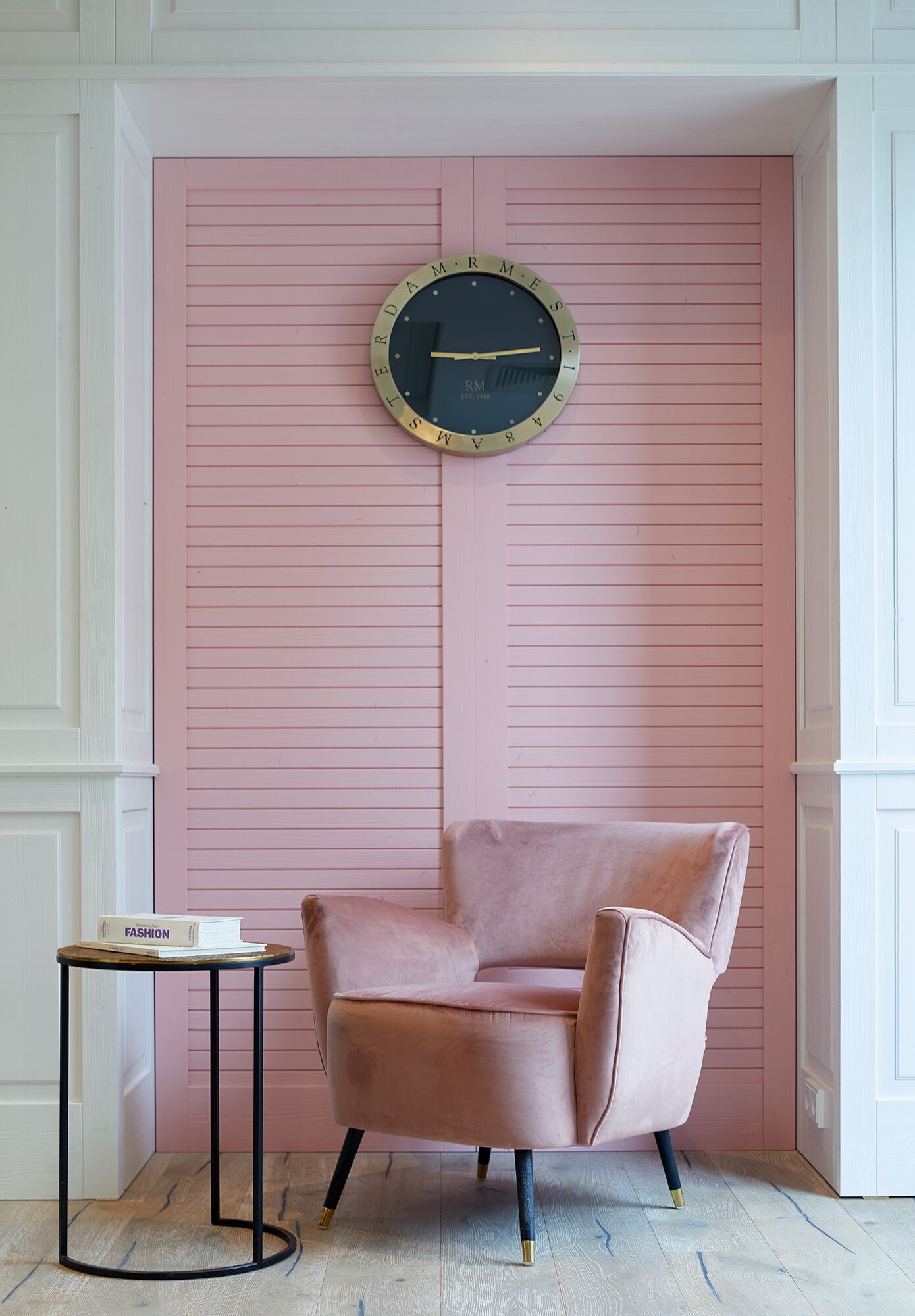 Een roze muur met een ronde klok, een roze stoel en een klein tafeltje met een boek.
