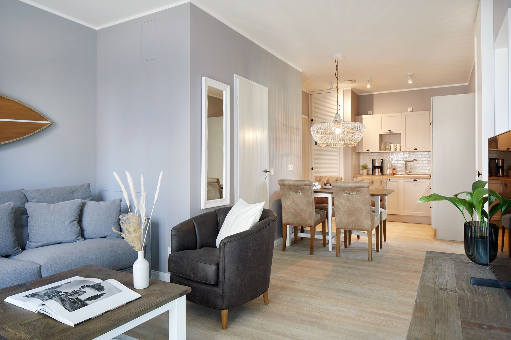 A modern living room with a grey sofa, a coffee table with a magazine, and a dining area with a chandelier.