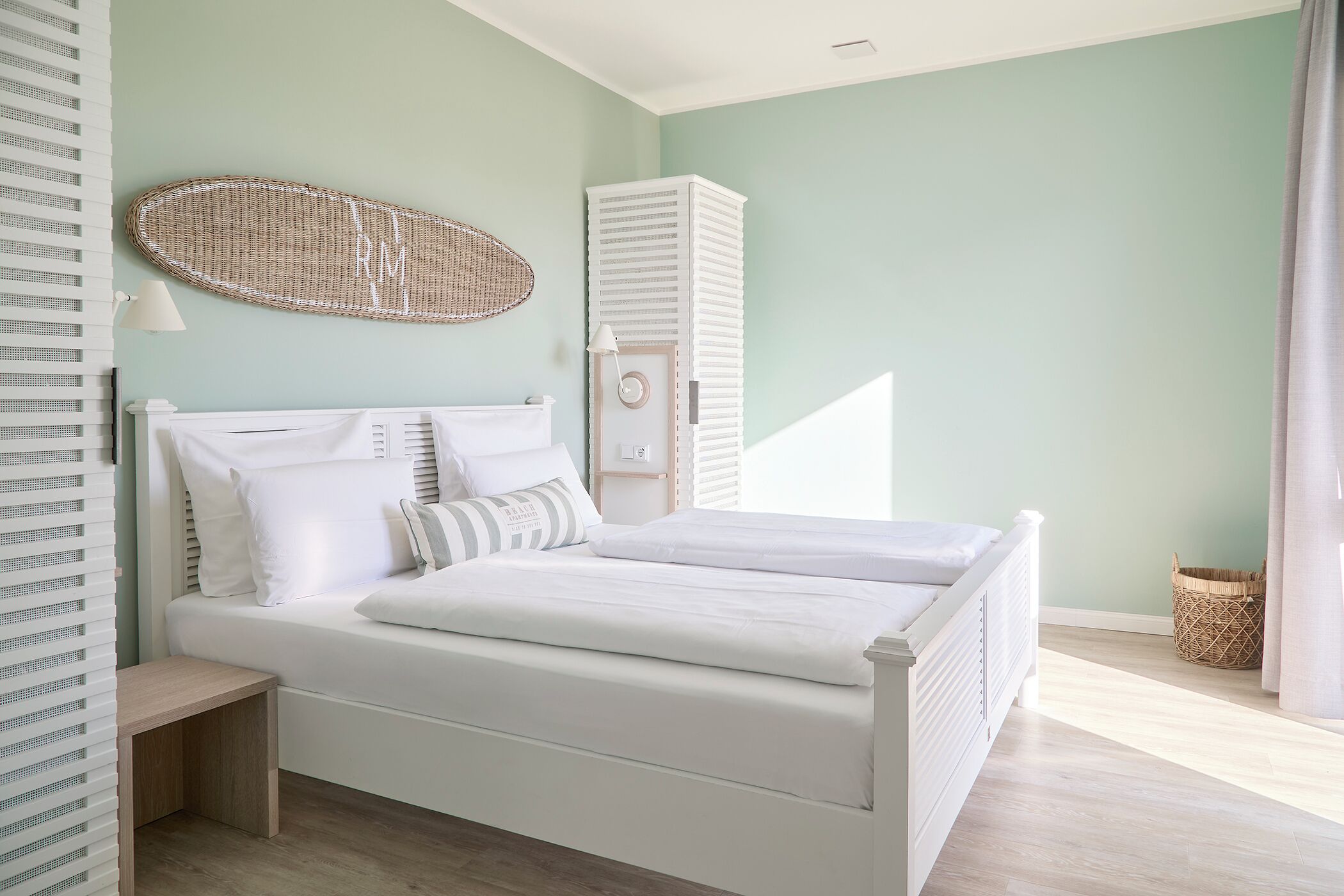 A neatly arranged bedroom with a bed, bedside tables, a wicker wall hanging, a tall white cabinet, and a wicker basket on the floor.