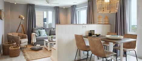 A modern living room with a sofa, a dining table with chairs, a small kitchen area, and a large window with curtains.