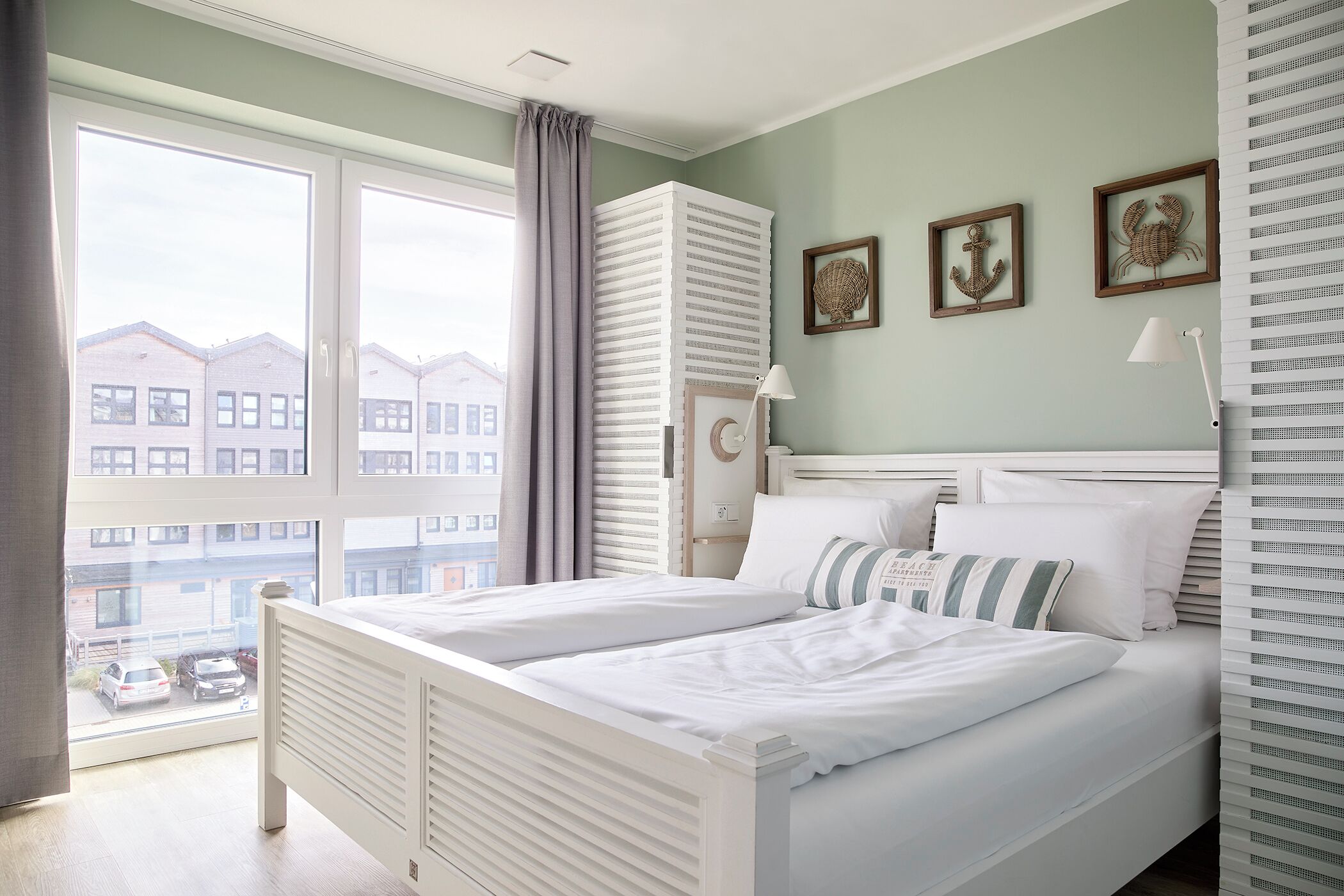 Een slaapkamer met een groot raam, een bed met witte lakens, een nachtkastje en een lamp. Er hangen afbeeldingen in lijsten aan de muur.
