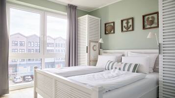 Een slaapkamer met een groot raam, een bed met witte lakens, een nachtkastje en een lamp. Er hangen afbeeldingen in lijsten aan de muur.