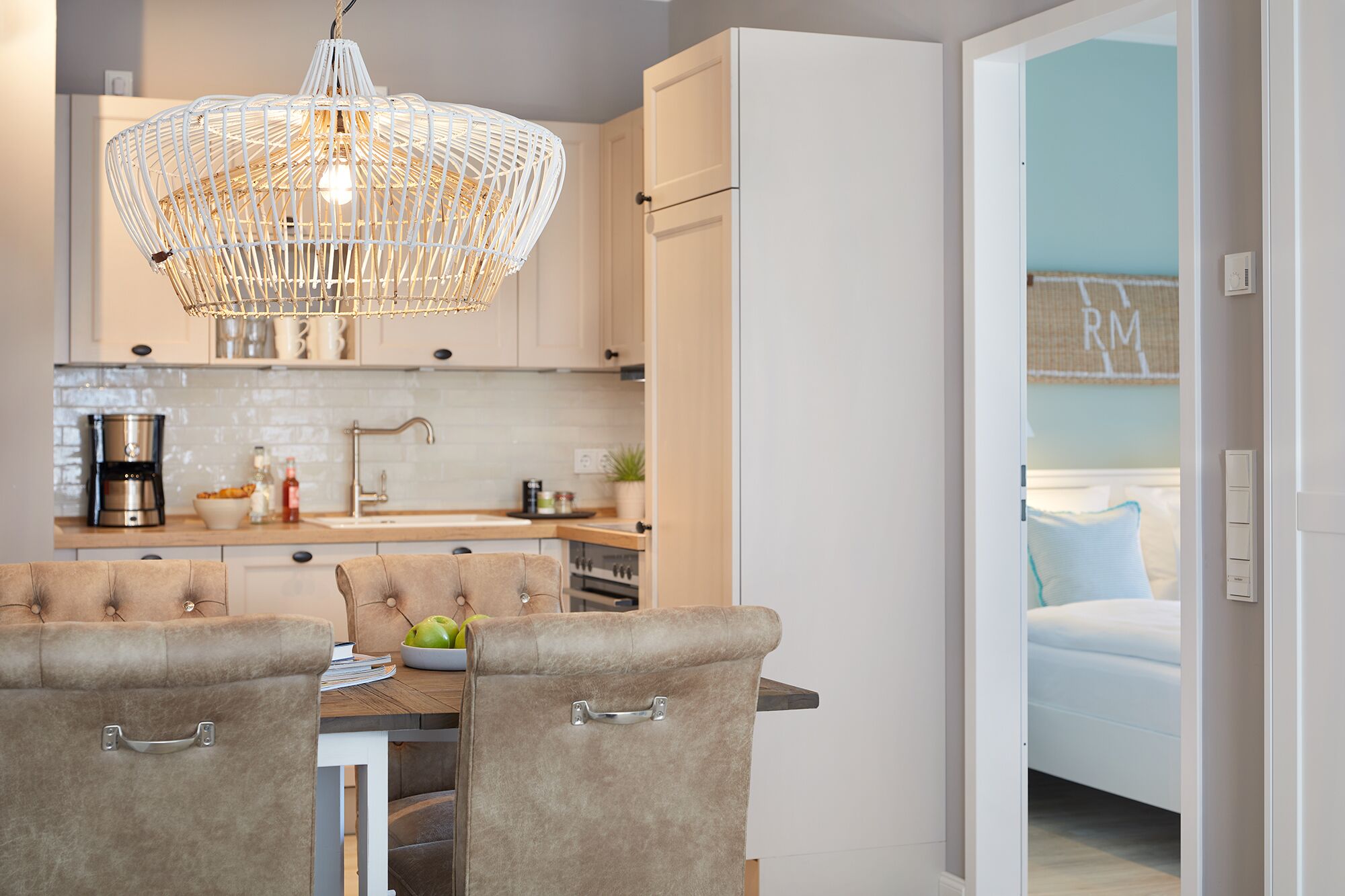 A modern kitchen with a dining area, featuring a chandelier, white cabinets, and a view into a bedroom.