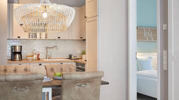 A modern kitchen with a dining area, featuring a chandelier, white cabinets, and a view into a bedroom.