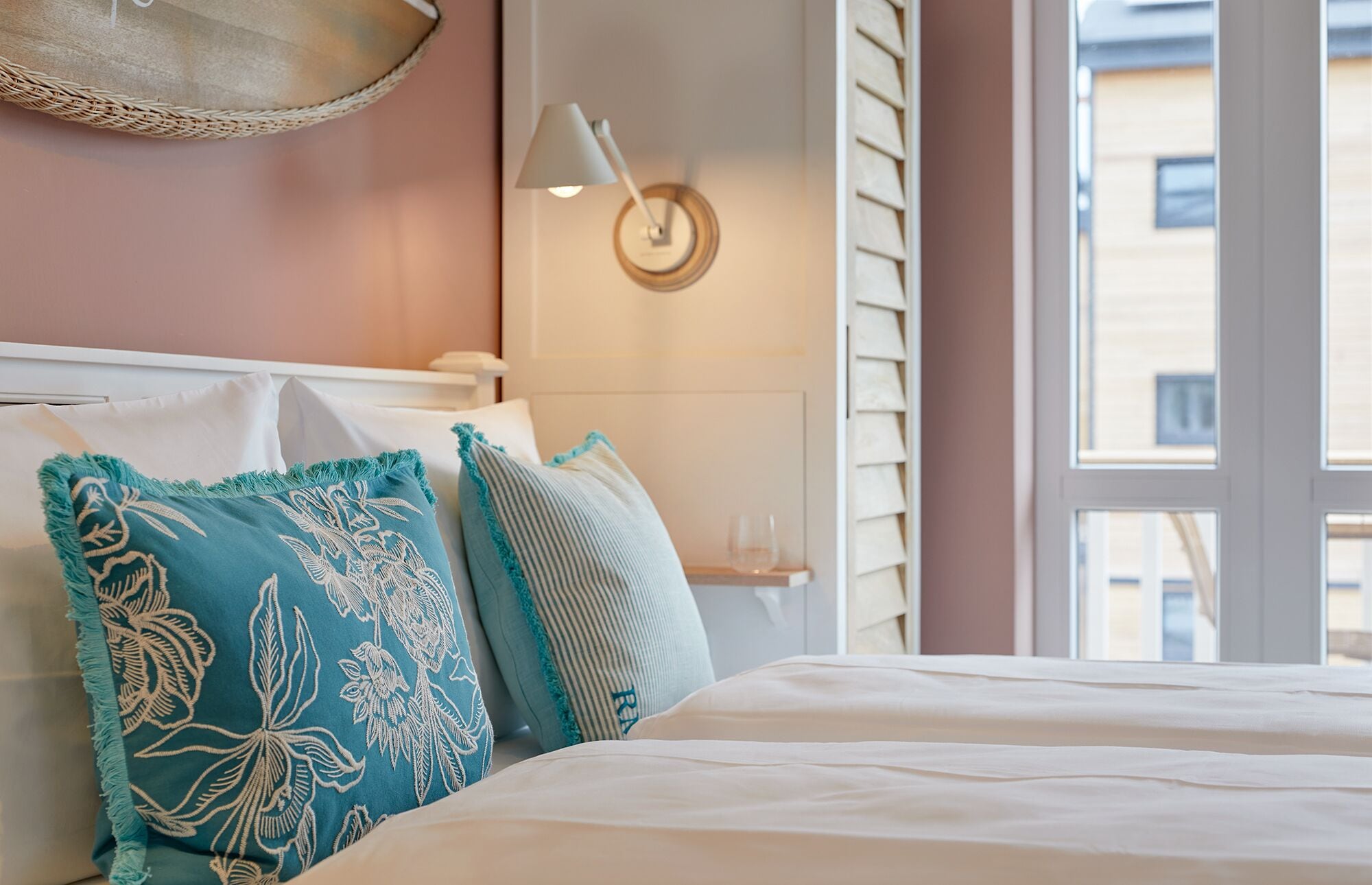 A neatly made bed with white bedding and decorative pillows, a bedside lamp, and a view of the outside through a window.