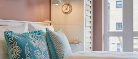 A neatly made bed with white bedding and decorative pillows, a bedside lamp, and a view of the outside through a window.