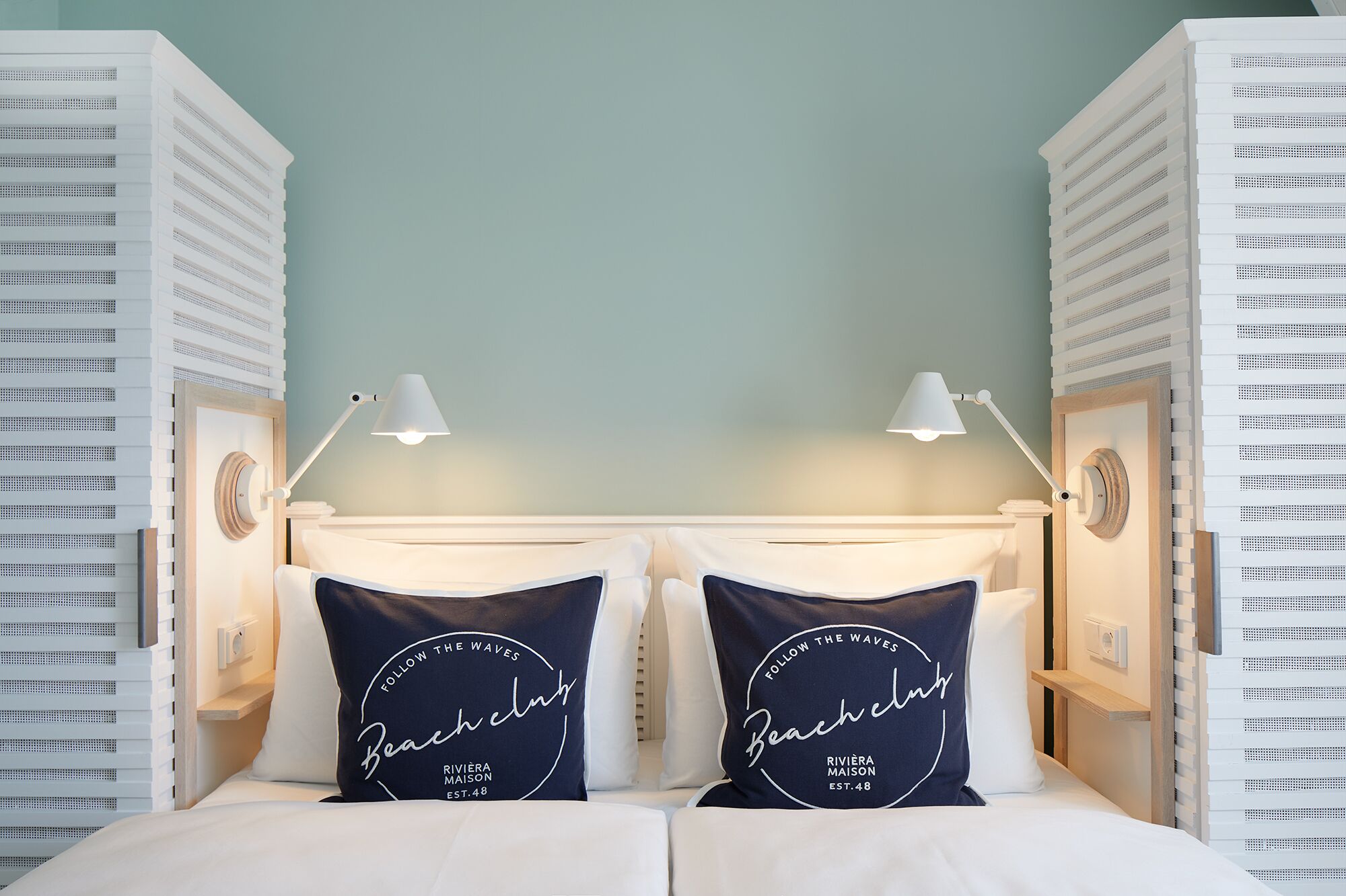 Une chambre d’hôtel avec deux lits, chacun agrémenté d’un oreiller orné des inscriptions « Follow the Waves Beach club » et « Rivièra Maison EST. 48 ».