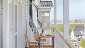 A balcony with two wicker chairs, a small table, and a view of a body of water and buildings in the distance. - Beach Apartments Büsum (Büsum)