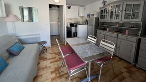 Un espace de vie compact comprenant un coin cuisine, un coin repas avec des chaises en métal, un canapé avec des coussins à motifs et une petite table.