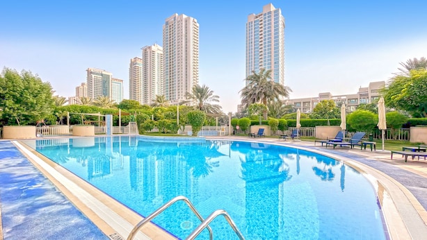 Una piscina con agua azul cristalina, rodeada de palmeras y edificios modernos al fondo.