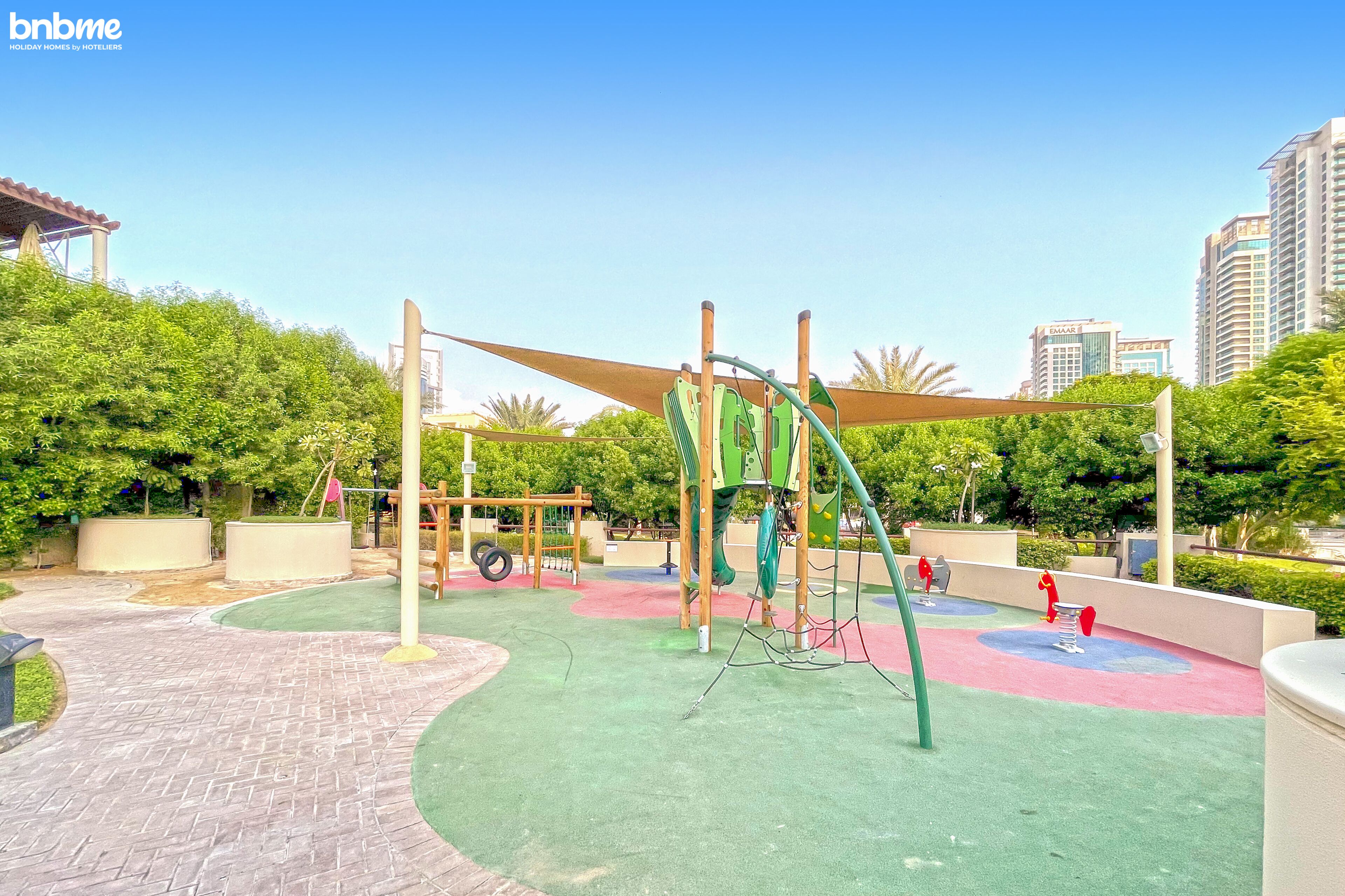 Un parque infantil con diversas estructuras de juego, rodeado de vegetación y una zona pavimentada.