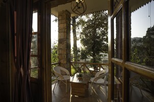 Un balcón abierto con una mesa de madera y dos sillas de mimbre blanco, con vista a un jardín y árboles.