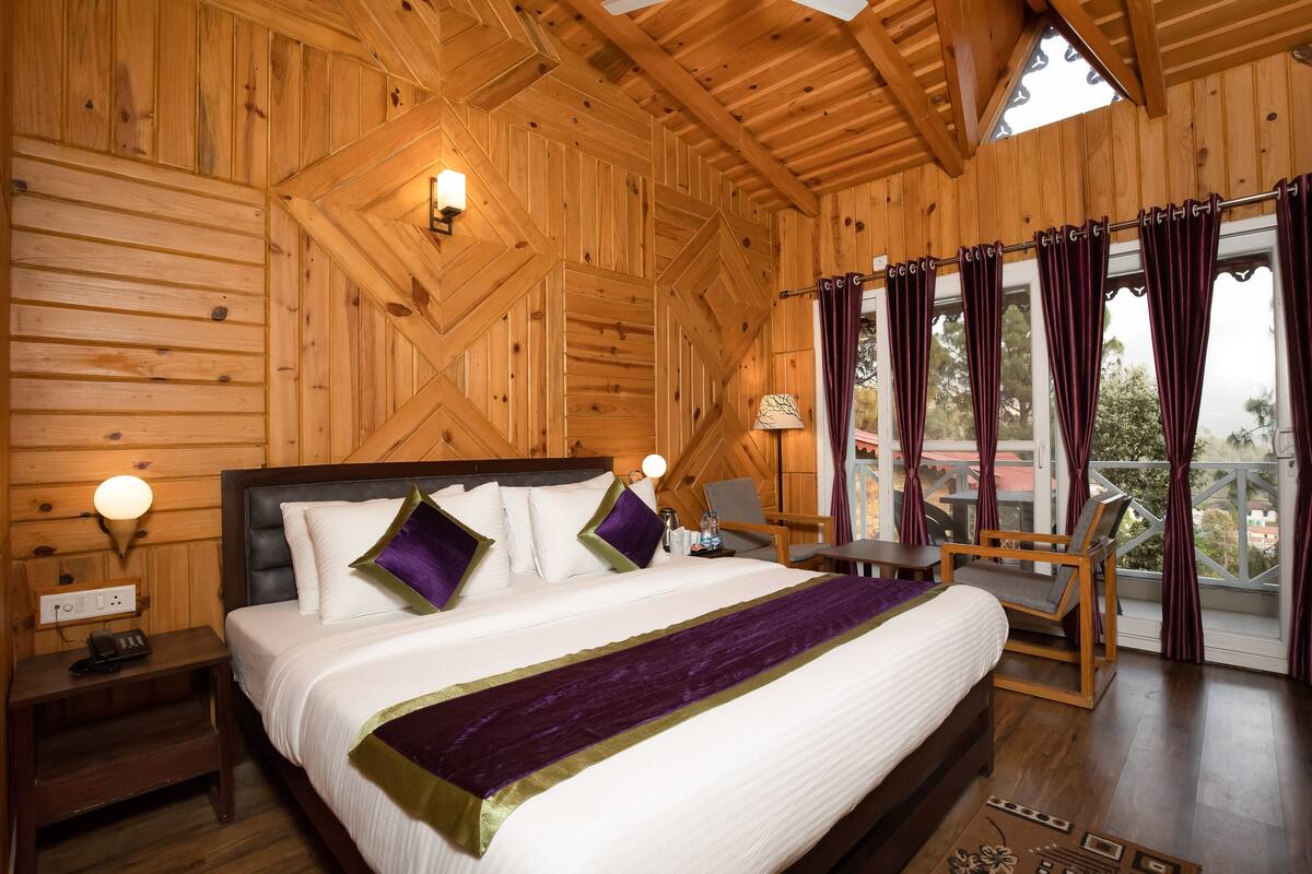 Un dormitorio tipo cabaña de madera con una cama grande, una mesita de noche, una silla y una ventana con cortinas.