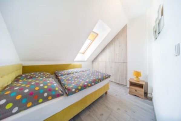Ein kleiner, heller Raum mit schrägem Dach und Oberlicht. Er verfügt über ein Bett mit farbenfroher, punktierter Bettwäsche und einen kleinen Nachttisch mit Lampe.