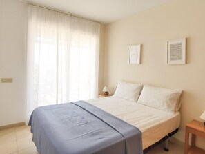 Ein Schlafzimmer mit Bett, Nachttisch, Lampe, einem Fenster mit Vorhängen und zwei gerahmten Bildern an der Wand.
