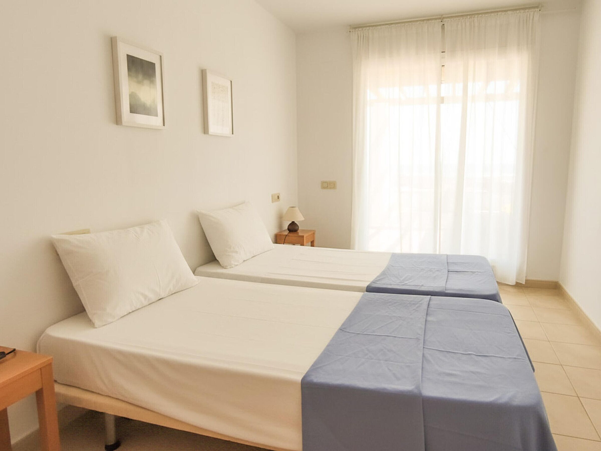 A room with two beds, a nightstand, a lamp, and a window with curtains.