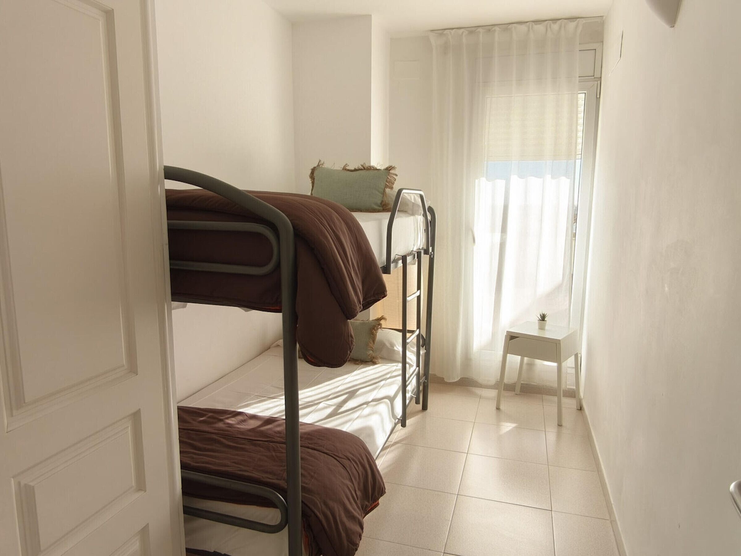 A dormitory room with bunk beds, a small table, and a window with sheer curtains.
