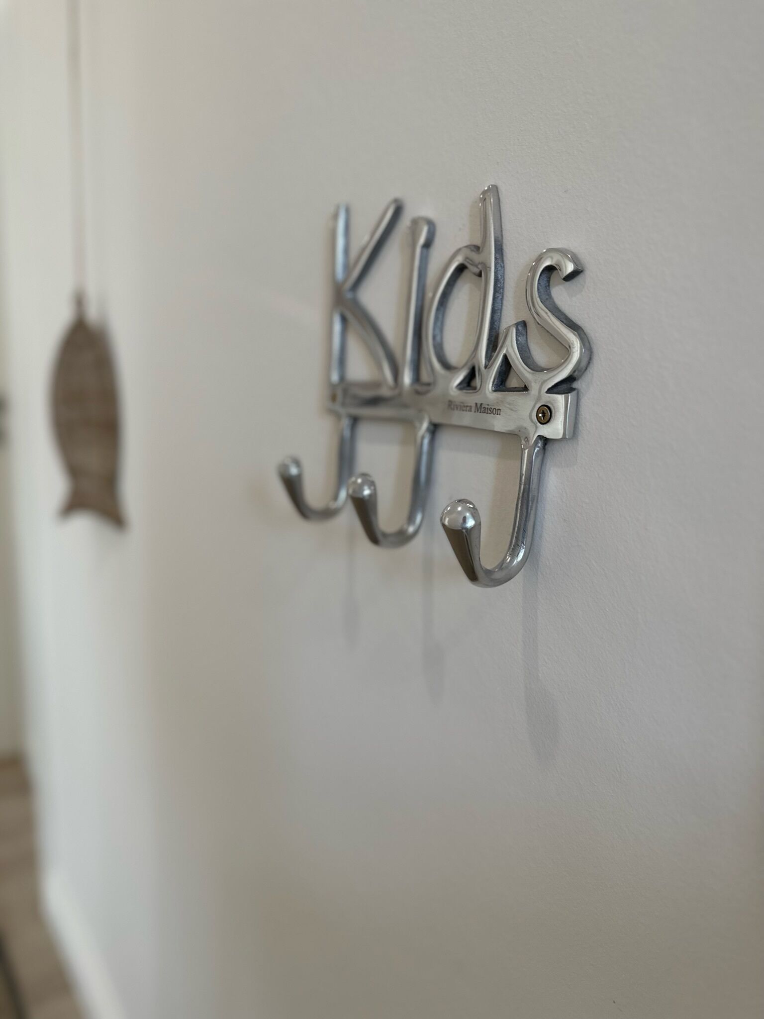 Un crochet métallique fixé au mur, portant le mot « Kids » et une petite plaque sur laquelle on peut lire « Riviera Maison ».