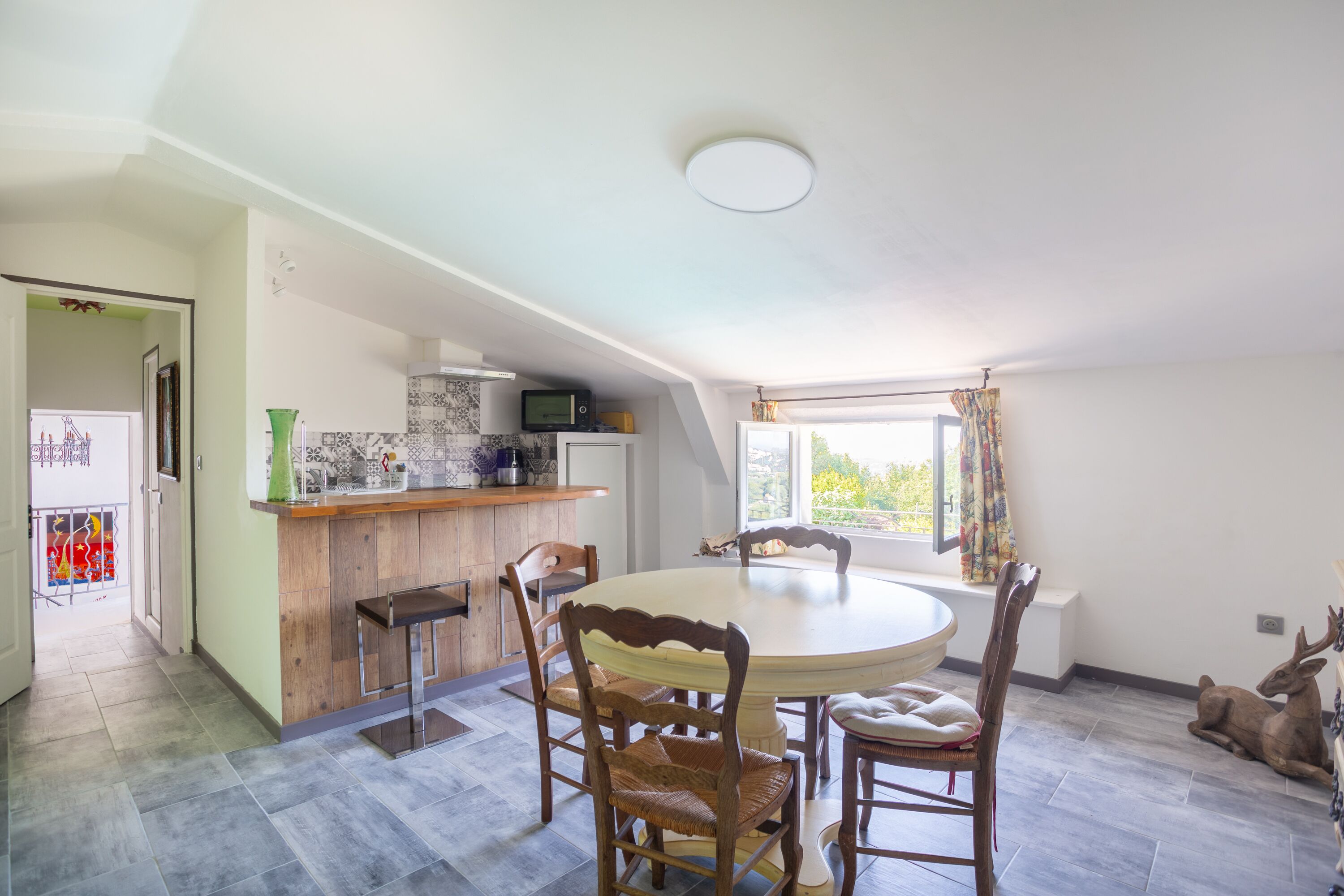 A dining area with a round table and wooden chairs, a kitchen with a microwave, and a window with curtains.