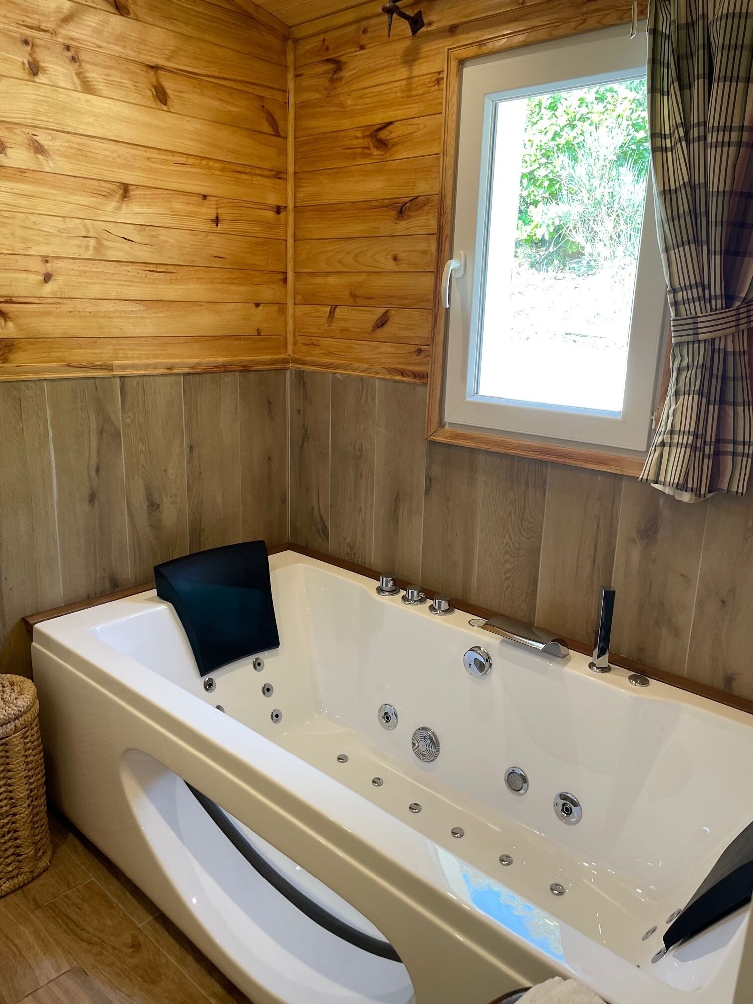 A wooden cabin interior with a large bathtub, a window with curtains, and a woven basket.