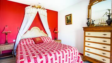 Een bed met hemel, een dressoir met lades, een nachtkastje met lamp en een lijst met een foto aan de muur.