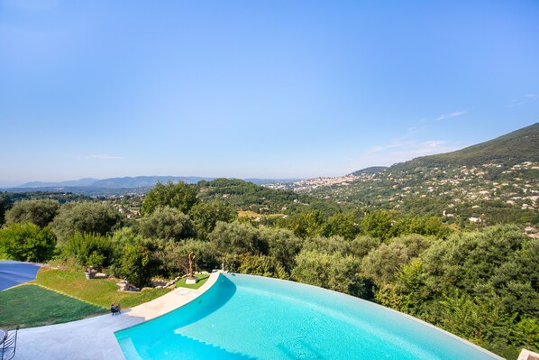 A swimming pool with a curved edge, surrounded by lush greenery and a view of distant hills and a town. - Le Clos des Senteurs (Chateauneuf-Grasse)