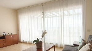 A living room with a sofa, a wooden coffee table, a cabinet, and a large window with sheer curtains. - Charming Apartment with Marina View in L'Ametlla de Mar (L'Ametlla de Mar)