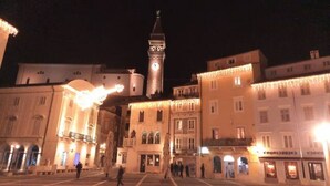 A square with a clock tower, illuminated buildings, and a few people walking.