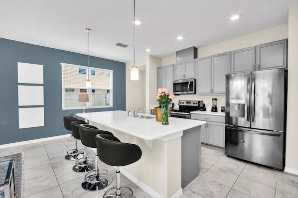 A modern kitchen with grey cabinets, stainless steel appliances, a central island with bar stools, and pendant lighting.