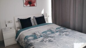 Una cama bien hecha con una colcha estampada, una mesita de noche blanca con una lámpara, un enchufe y un interruptor de luz en la pared.