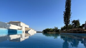 A clear blue infinity pool with a tiled floor, surrounded by a white building with a tiled roof, and a tall tree on the right. - SOLAR ALVURA HEALTH HOTEL (Olhao)