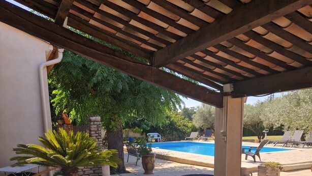Una vasca idromassaggio sotto un pergolato di legno, con una piscina e sedie a sdraio sullo sfondo.