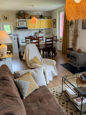 Una sala acogedora con un sofá marrón, una zona de comedor con sillas de madera y una mesa, una cocina con armarios amarillos y una lámpara naranja colgante, y una chimenea.