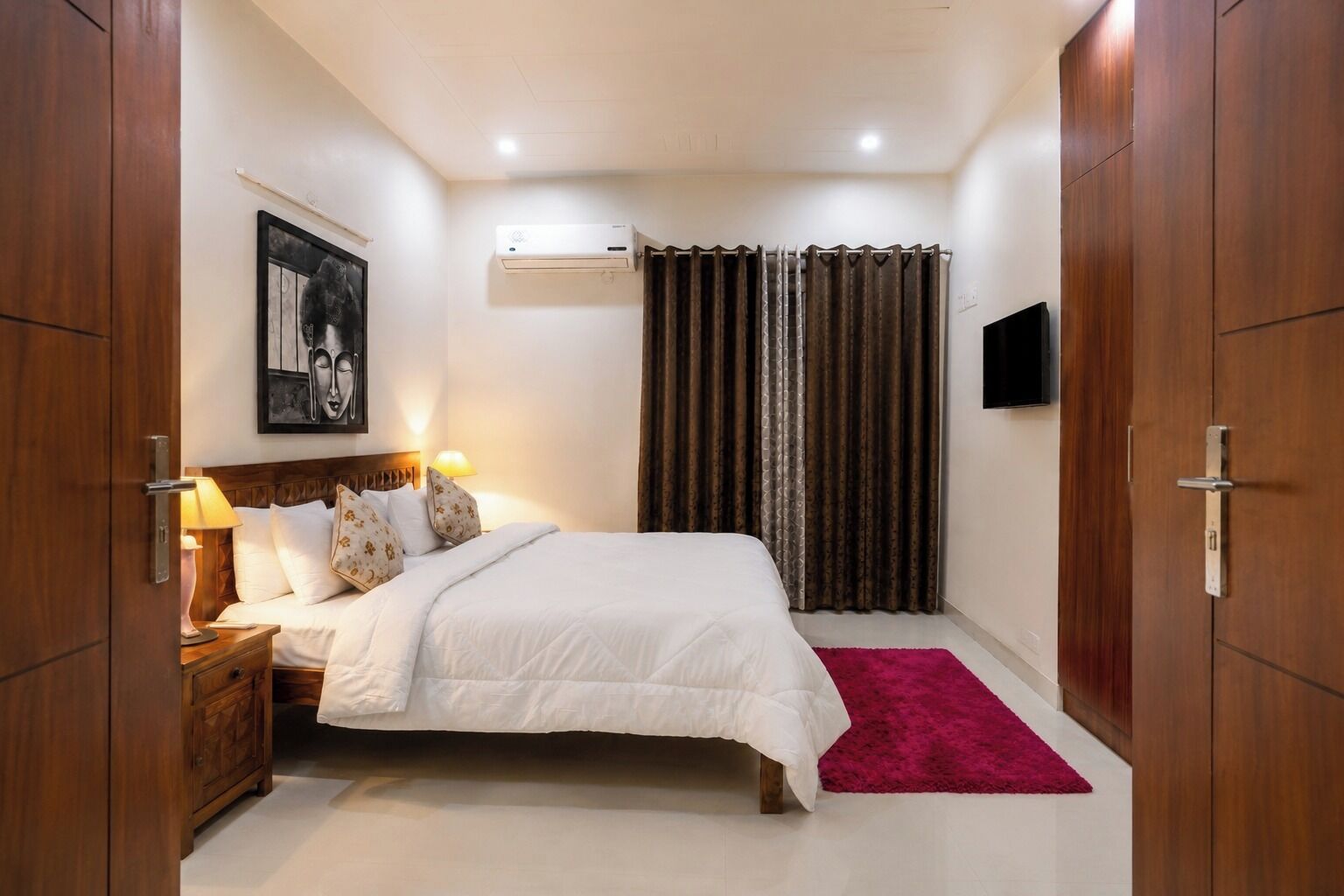 A bedroom with a bed, bedside tables, a TV, a wall-mounted air conditioner, and a window with curtains.