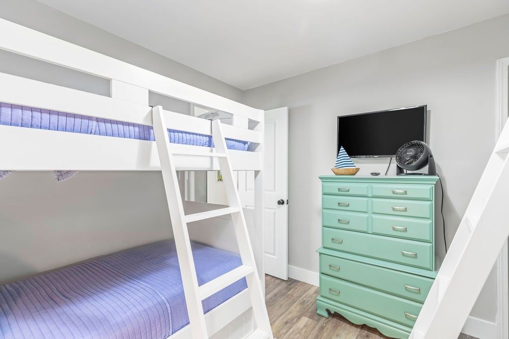 A child's bunk bed with a ladder, a television, and a dresser in a room with a door and a window.