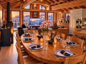 Un coin repas avec une table en bois dressée pour le repas, entourée de chaises, avec une cheminée et une vue panoramique sur les montagnes par les fenêtres.