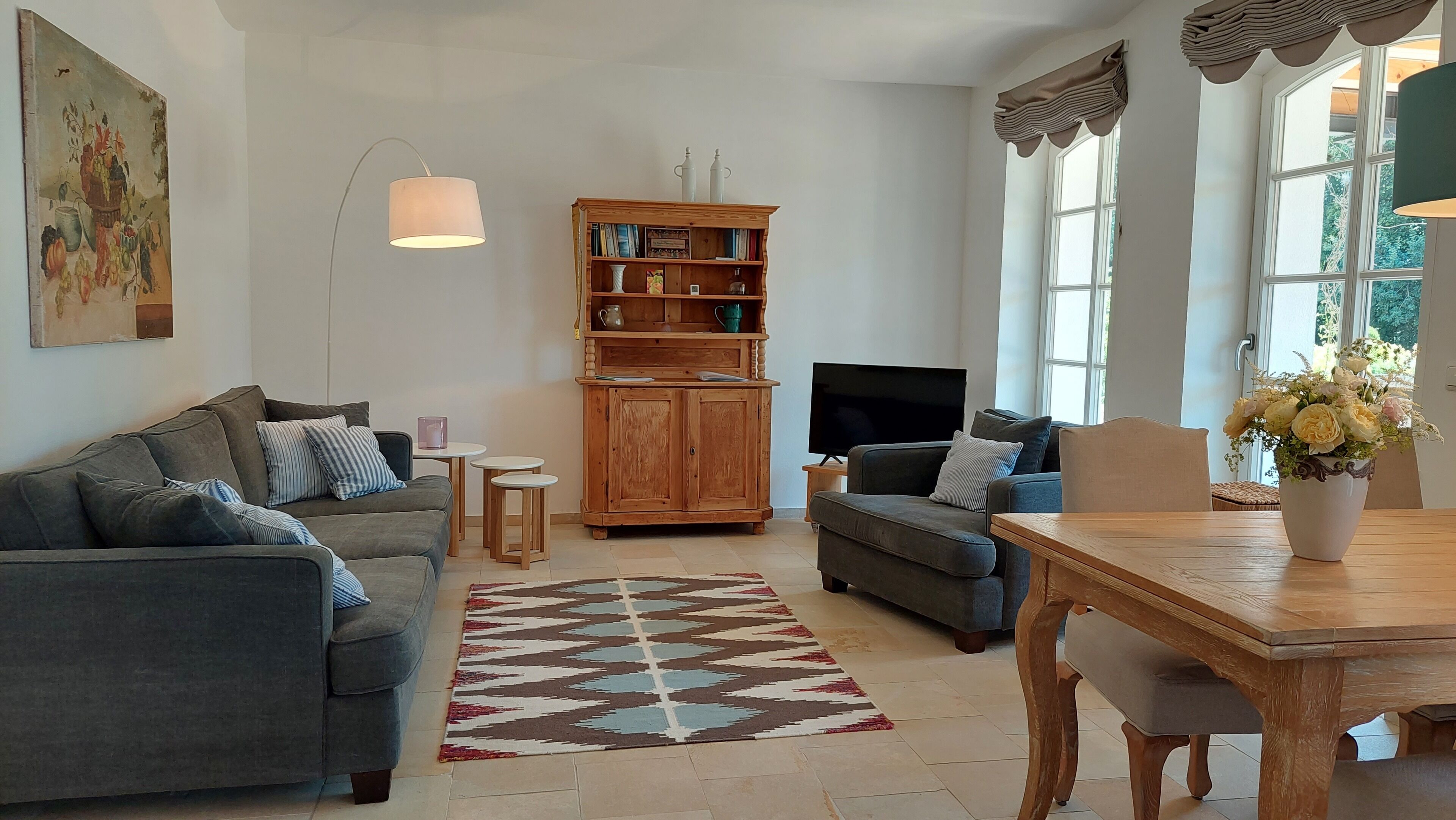 A living room with a grey sofa, a smaller grey armchair, a wooden cabinet, a TV, and a dining table with a vase of flowers.