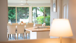 A modern kitchen with a central island, a dining area with chairs, and large windows offering a view of greenery.