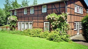 Un bâtiment à deux étages doté d’une façade traditionnelle à pans de bois, de nombreuses fenêtres et d’un jardin bien entretenu où poussent diverses plantes et fleurs.