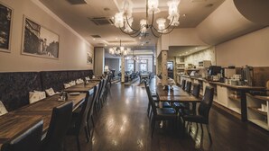 Un restaurant spacieux avec de longues tables et des chaises en bois, un lustre central et une vue sur la rue grâce à de grandes fenêtres.