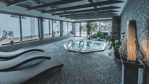 Un espace piscine intérieur moderne offrant une vue dégagée sur l’extérieur, un coin salon avec des chaises longues et un mur en pierre au motif texturé.