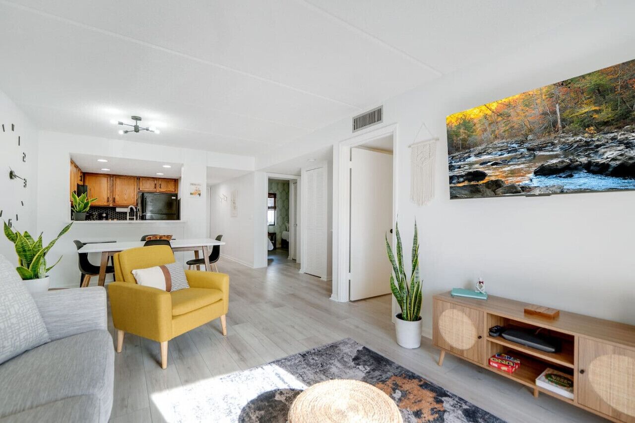 Una sala moderna con un sofá, un sillón amarillo y un comedor con mesa y sillas. Se ve una cocina al fondo, y un cuadro de un río en la pared.