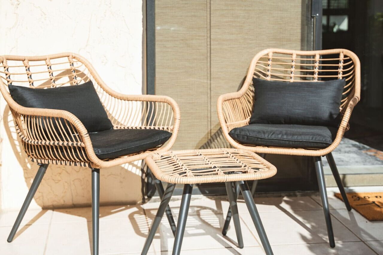 Twee rieten stoelen met zwarte kussens en een bijpassend voetenbankje op een betegelde vloer.